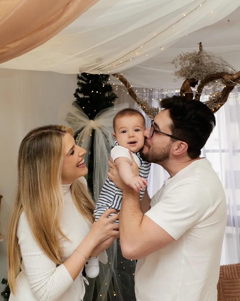 Casal com o bebê entre os dois, o pai beijando o rosto do bebê, no cenário de Natal do estúdio com tule e luzes