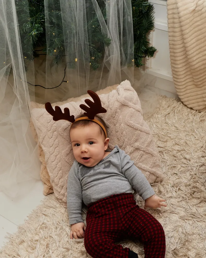 Bebê deitado em almofadas e tapete macio, com chifres de rena, no cenário de Natal do estúdio