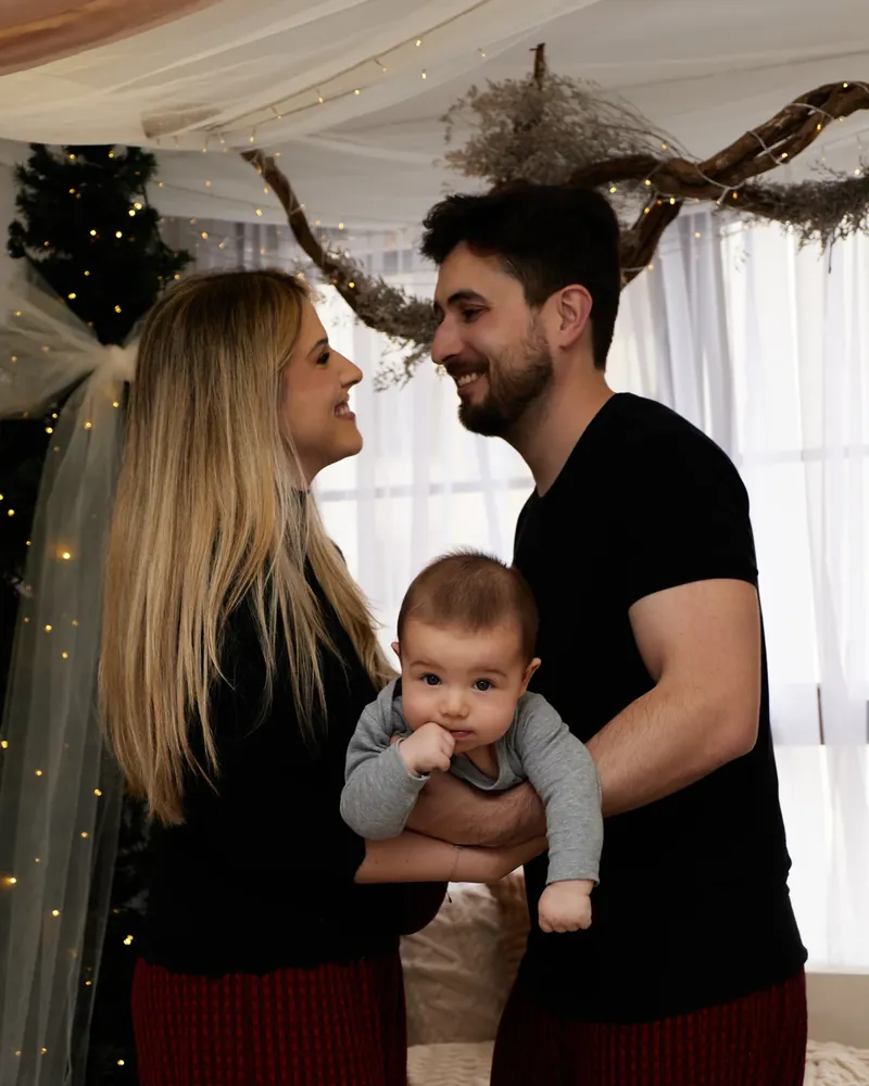 Casal olhando-se com o bebê entre eles, no cenário de Natal do estúdio com árvore, tule e luzes