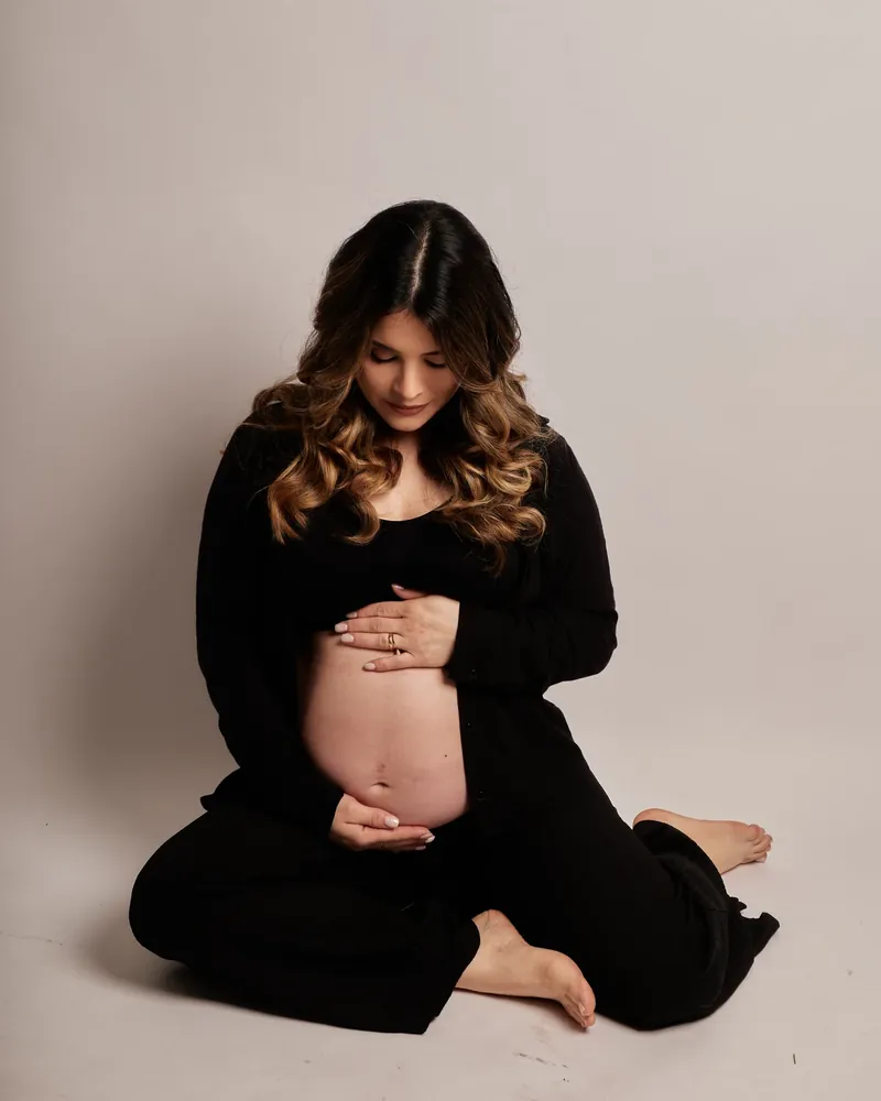Gestante sentada no chão, de roupa preta, com as mãos na barriga, em ensaio de gestante no estúdio