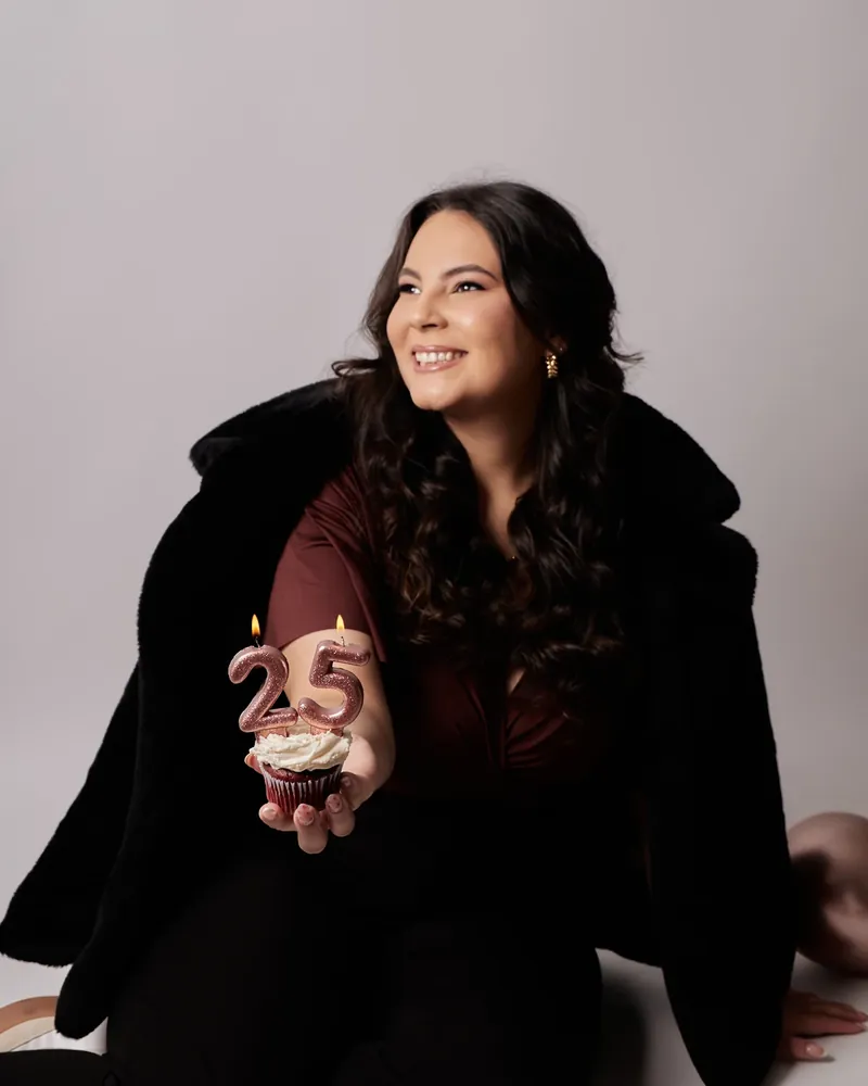 Mulher sorrindo, com casaco preto de pelos, segurando um cupcake com vela em forma do número 25