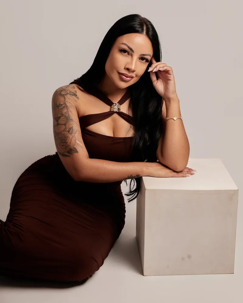 Mulher de cabelo preto e vestido marrom sentada ao lado de um cubo branco, em ensaio feminino no estúdio